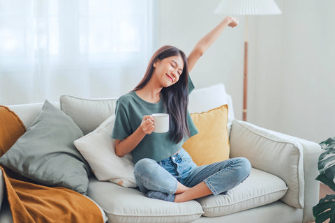 Frau sitzt auf Sofa mit Kaffee in der Hand und streckt einen Arm in die Luft