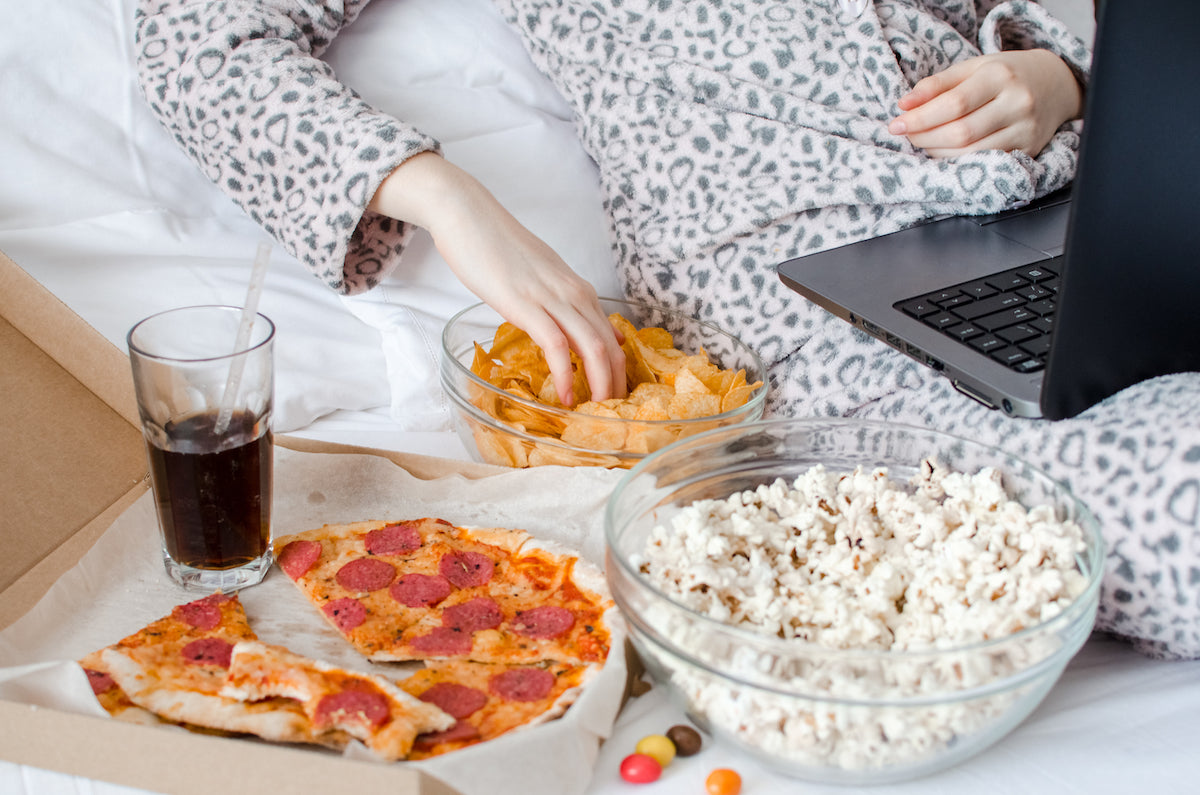 Gesunde Snacks für abends: Person liegt mit Laptop im Bett, daneben Pizza, Chips, Popcorn und Cola