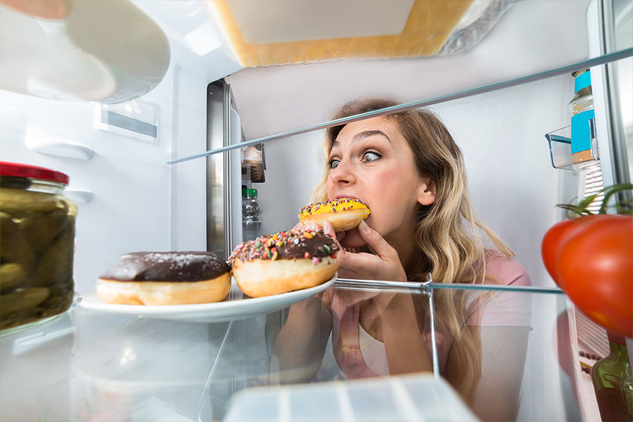 eine Frau nascht einen Donut aus einem Kühlschrank